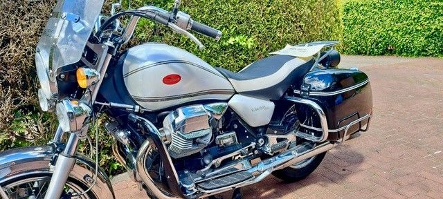 Ein silber-schwarzes Moto Guzzi-Motorrad, geparkt auf einer Ziegelfläche, im Hintergrund eine grüne Hecke.