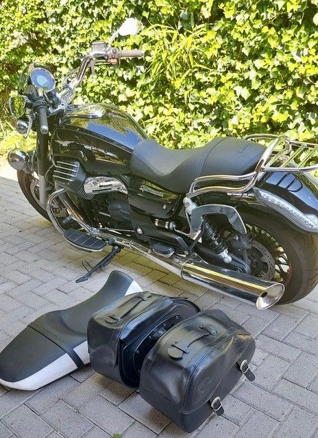 Schwarzes Motorrad mit Satteltaschen und Sitz auf einer Ziegelfläche vor Grün.