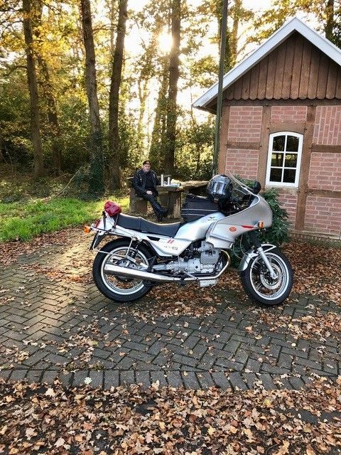 Ein silbernes Motorrad parkte auf einem Kopfsteinpflasterweg neben einem Backsteingebäude in einem Waldgebiet.