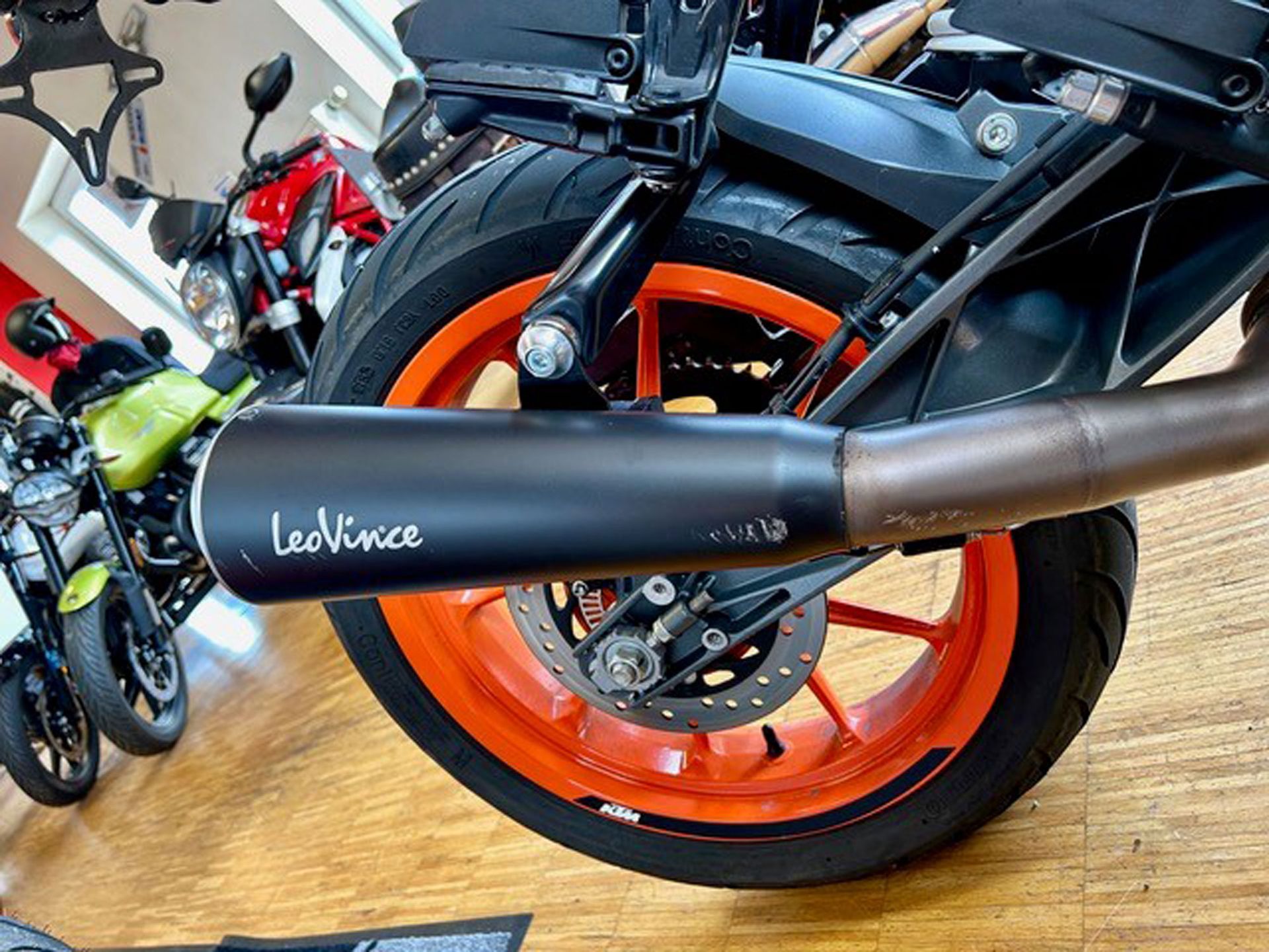 Nahaufnahme eines schwarzen LeoVince-Motorradauspuffs an einer KTM Duke mit orangefarbenen Rädern. Im Hintergrund sind andere Motorräder zu sehen.