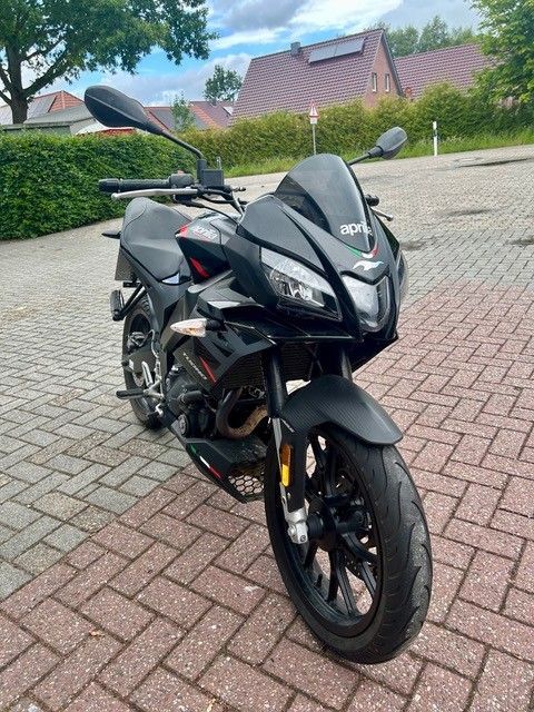 Ein schwarzes Sportmotorrad parkt auf einer gepflasterten Auffahrt; im Hintergrund sind Häuser und Hecken zu sehen.
