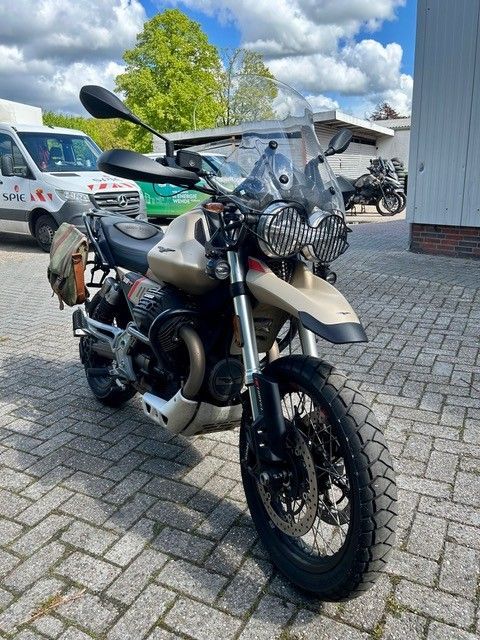Ein Adventure-Motorrad parkt auf Kopfsteinpflaster vor einem Gebäude; es ist mit einer Fronttasche und Spiegeln ausgestattet.