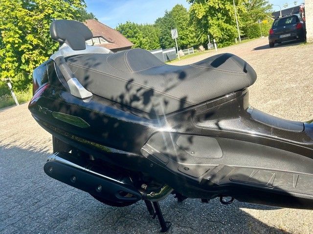 Schwarzer Motorroller mit hochgeklapptem Sitz und sichtbaren Beschädigungen parkt in einer Wohnstraße.
