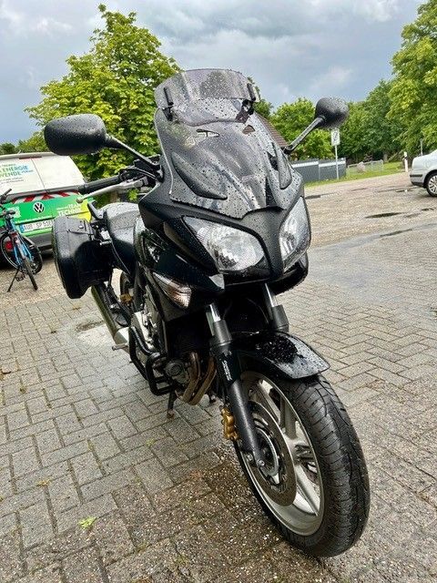 Schwarzes Tourenmotorrad parkt auf einem nassen, asphaltierten Parkplatz; im Hintergrund Bäume und bewölkter Himmel.