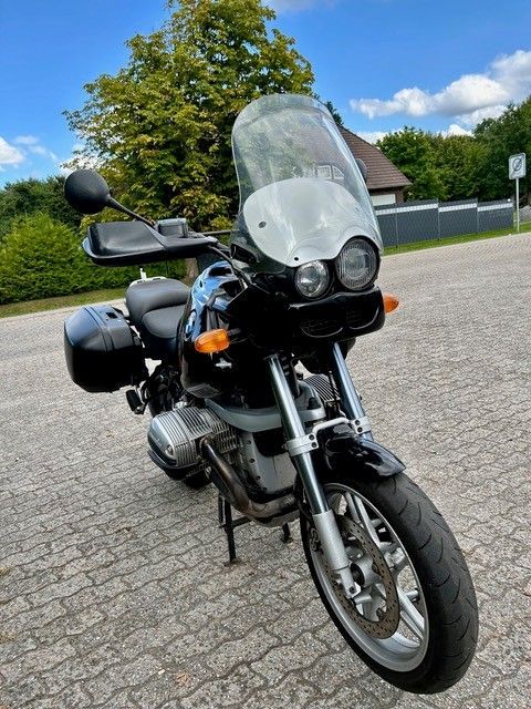 Schwarzes BMW-Motorrad mit Windschutzscheibe, Seitentaschen und schwarzem Lenker, geparkt auf einer gepflasterten Fläche.