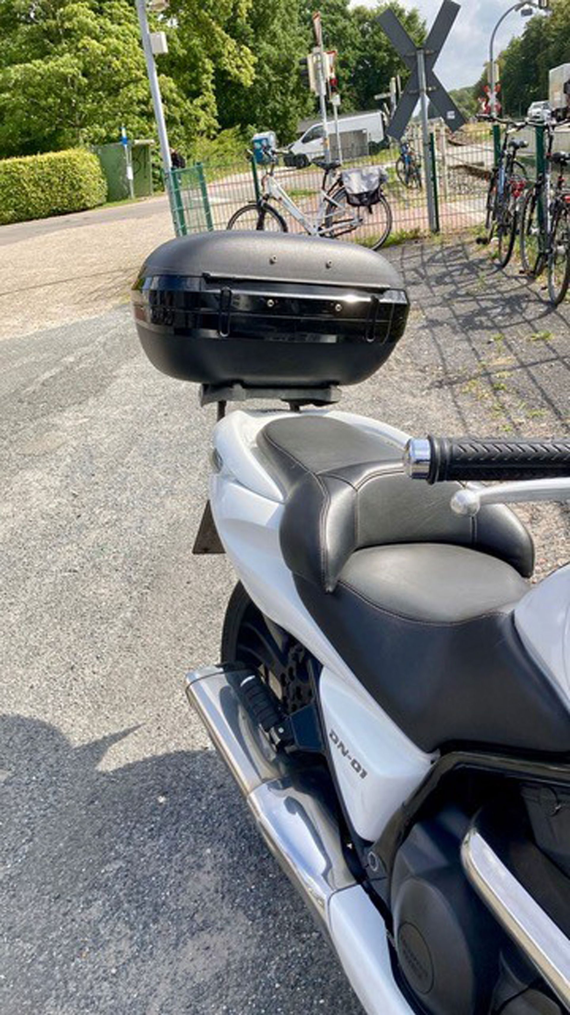 Weißer Motorroller mit schwarzem Topcase auf Schotterweg geparkt; Fahrräder und Grün im Hintergrund.