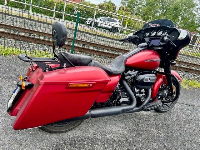 Ein rotes Tourenmotorrad mit roten Satteltaschen und schwarzer Windschutzscheibe parkt auf dem Bürgersteig neben Bahngleisen.