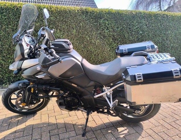 Grau-schwarzes Adventure-Motorrad mit Gepäck, geparkt auf einer gemauerten Auffahrt neben einer grünen Hecke.