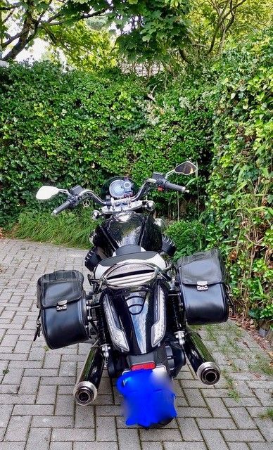 Schwarzes Motorrad mit Satteltaschen, geparkt auf einem Ziegelweg, umgeben von grünem Laub.