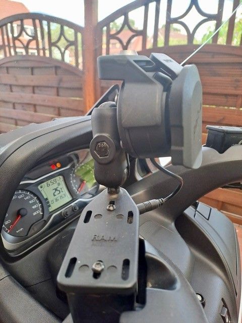 Nahaufnahme des Lenkers eines Motorrads mit einer Telefonhalterung, einem Tachometer und einem grauen Gerät. Das Motorrad ist im Freien geparkt.