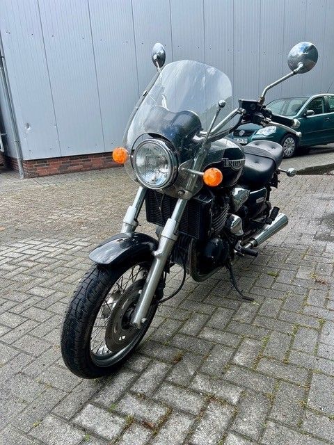 Schwarzes Motorrad mit Windschutzscheibe und Seitenspiegeln parkt auf einer gepflasterten Fläche vor einer grauen Mauer.