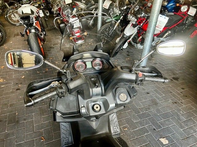 Motorradcockpit mit Lenker und Spiegeln auf einem Parkplatz, dahinter mehrere Roller.