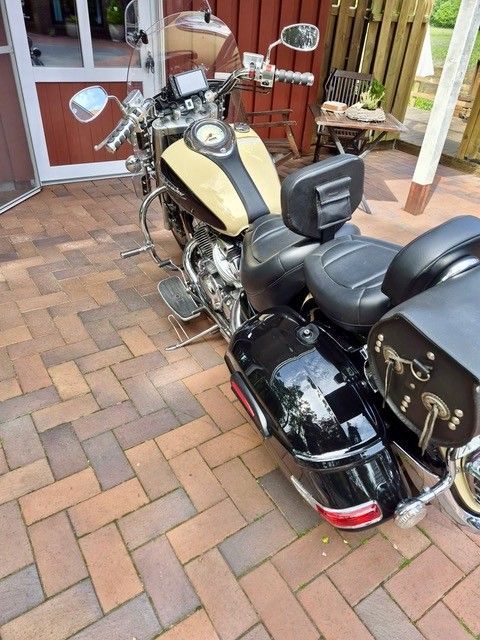Schwarzes Tourenmotorrad mit beigem Tank parkt auf einer Backsteinterrasse in der Nähe eines Hauses