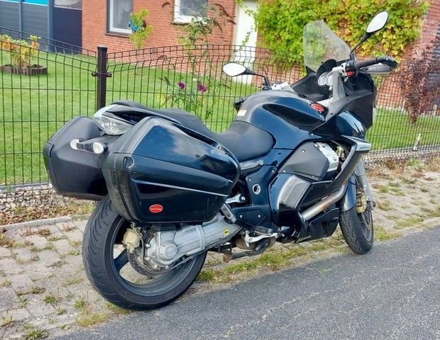 Ein dunkelblaues Moto Guzzi-Motorrad mit Packtaschen, das auf einer gepflasterten Fläche in der Nähe eines Zauns geparkt ist.