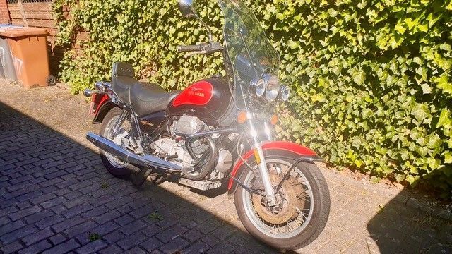 Ein rot-silbernes Motorrad parkt im Sonnenlicht vor einer Hecke.