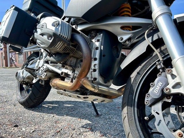 Im Freien auf dem Asphalt sind Motor, Auspuff und Vorderreifen eines silber-schwarzen BMW-Motorrads zu sehen.