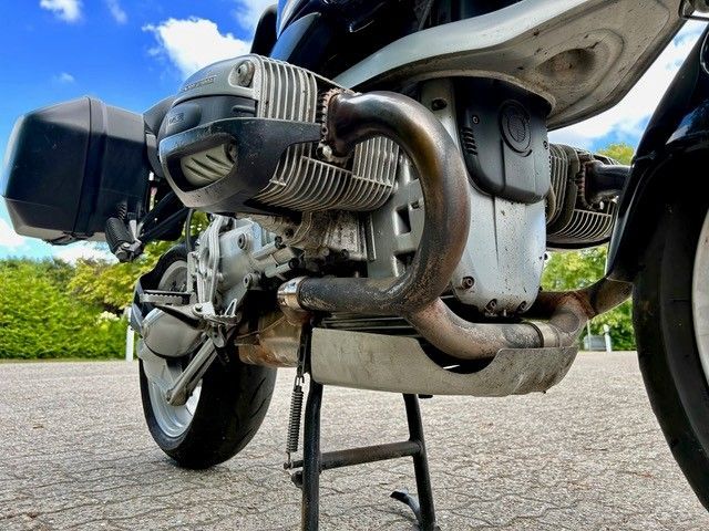 Nahaufnahme des Motorradmotors, Silber, Schwarz und Metallteile sichtbar. Bodenansicht, auf einem Ständer geparkt.