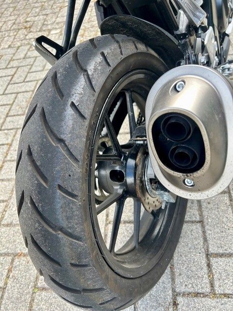Hinterrad und Auspuff eines geparkten Motorrads auf einem gepflasterten Gehweg.