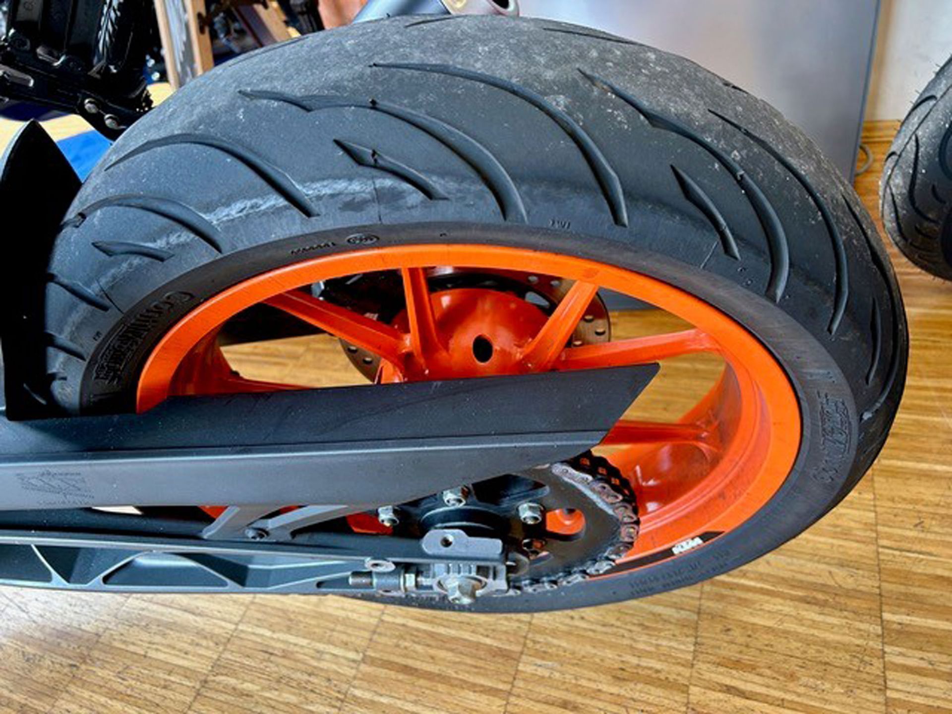 Hinterrad eines orange-schwarzen Motorrads mit Reifen, Felge, Kette und Schwinge, Innenaufnahme.