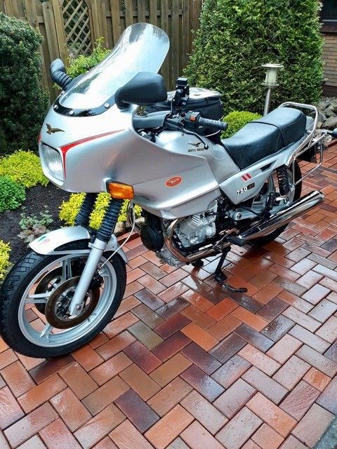 Ein silbernes Sportmotorrad parkt auf einer gemauerten Terrasse neben einem Holzzaun und grünen Sträuchern.