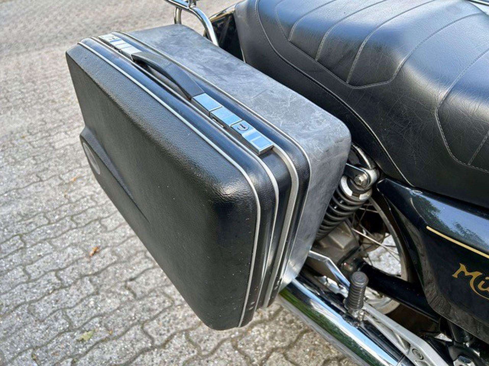 Ein schwarzer Koffer, der als Seitentasche an einem Motorrad befestigt wird. Der Koffer hat eine silberne Verzierung und wird seitlich in der Nähe des Sitzes montiert.