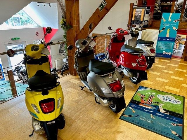 Mehrere Motorroller in einem Ausstellungsraum, darunter in Gelb, Silber und Rot. Holzbalken und Holzboden.