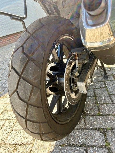Nahaufnahme eines Roller-Vorderrads und der Bremse auf einem Ziegelpflaster, zu sehen sind ein Reifen mit dickem Profil und eine schwarze Felge.