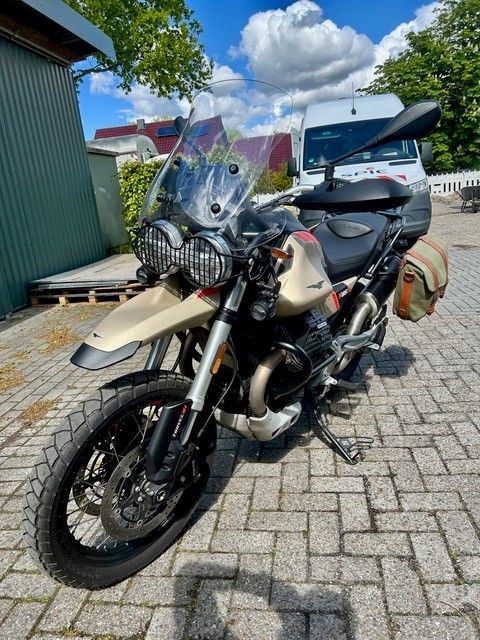 Ein beiges Adventure-Motorrad parkt auf einer gepflasterten Auffahrt neben einem grauen Auto und einem blauen Haus unter bewölktem Himmel