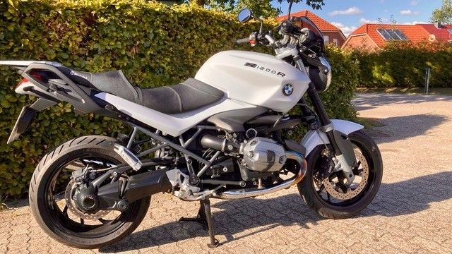 Weißes Motorrad BMW R1200R, geparkt auf einer gepflasterten Fläche, mit einer Hecke und Gebäuden im Hintergrund.