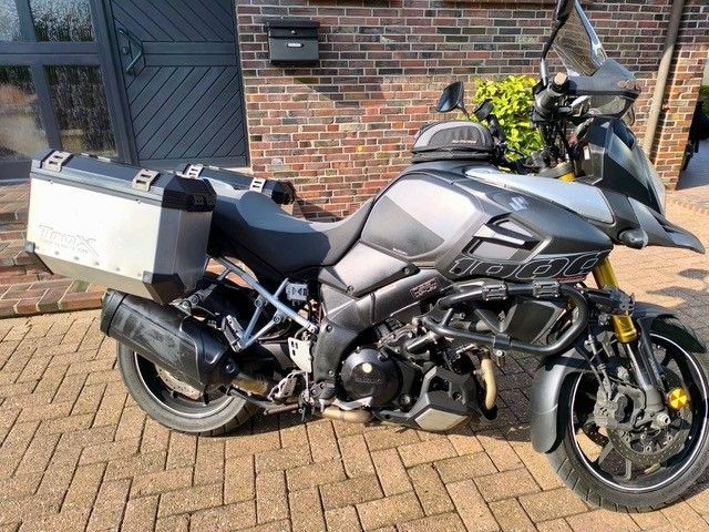 Ein graues Suzuki V-Strom-Motorrad mit harten Seitenkoffern, geparkt auf einem Ziegelpflaster vor einem Backsteingebäude.