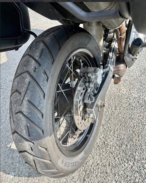 Hinterreifen eines Motorrads, Schrägansicht. Schwarzer Reifen mit Profil auf einem Speichenrad. Bremse und Auspuff sind ebenfalls sichtbar.