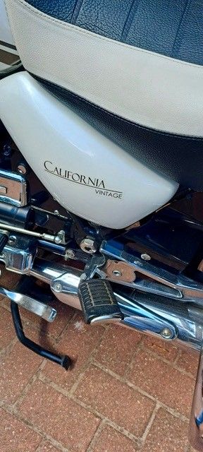 Motorrad mit Aufschrift „California“. Sitzfläche weiß und blau. Fußstütze und Auspuff sichtbar.