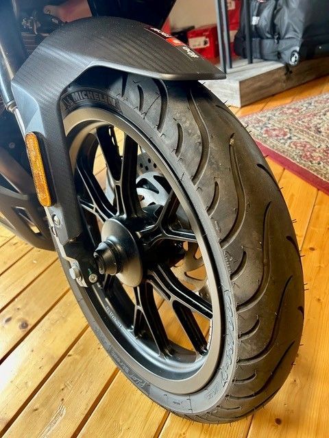Nahaufnahme eines Roller-Vorderrads mit schwarzer Leichtmetallfelge, der auf einem Holzboden in einem Innenraum abgestellt ist.