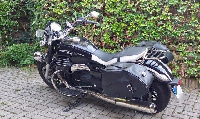 Schwarzes Moto Guzzi-Motorrad mit silbernen Akzenten, geparkt auf einer gepflasterten Auffahrt vor grünem Laub.