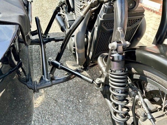 Vorderradaufhängung eines schwarzen Trikes. A-Arme, Stoßdämpfer und Rad sichtbar. Grauer Asphalt.