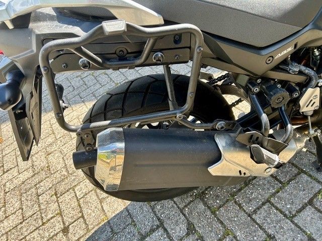 Motorrad-Seitenansicht mit schwarzem und verchromtem Auspuff und Gepäckträger, geparkt auf einem Ziegelpflaster.