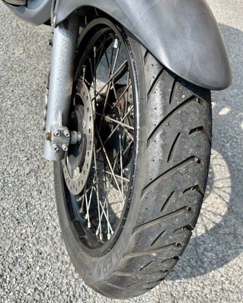 Nahaufnahme des Vorderrads eines Motorrads. Der Reifen ist schwarz mit abgenutztem Profil und die Speichen sind sichtbar.