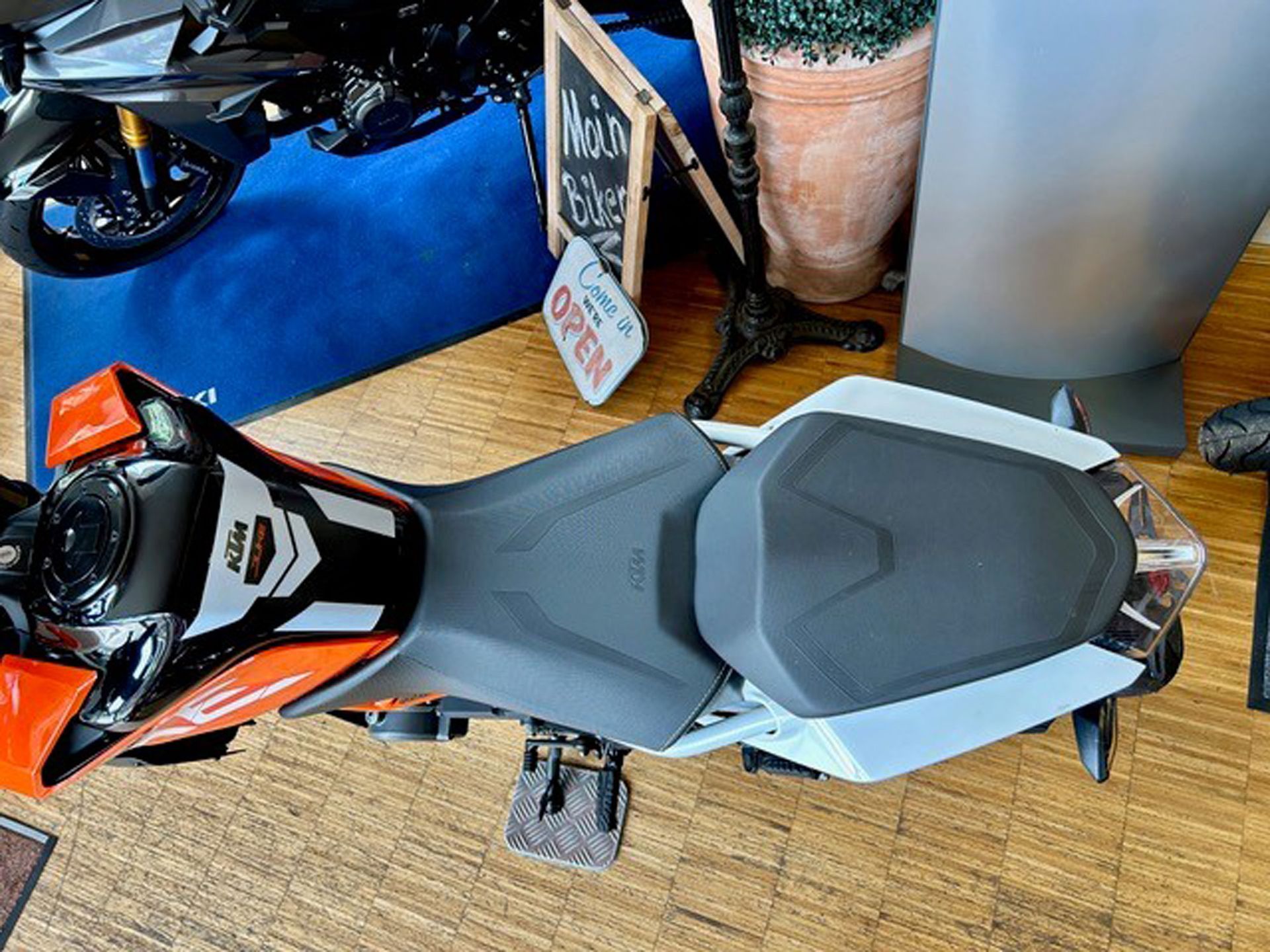 Orange-schwarzer Motorradsitz. Teilansicht mit einem „Geöffnet“-Schild im Hintergrund.