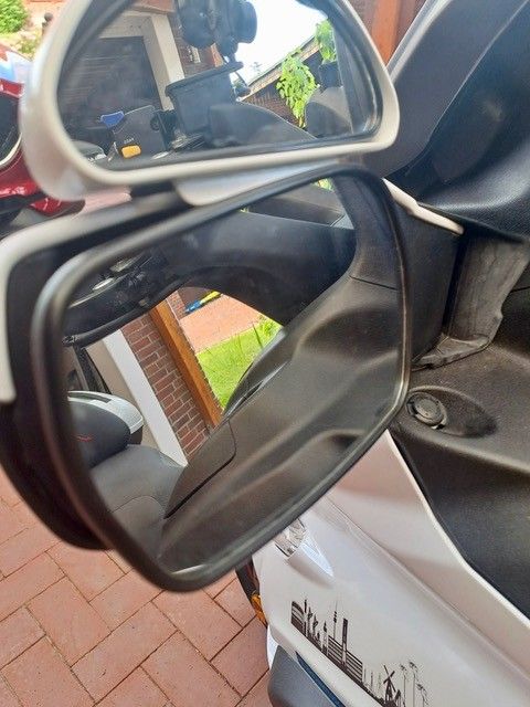 Nahaufnahme der Seitenspiegel eines Motorrollers, die den Blick auf den Lenker, die Handschuhe des Fahrers und die Umgebung ermöglicht.