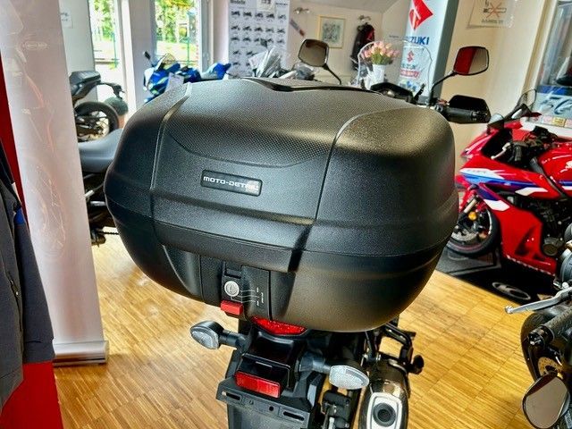Schwarzes Motorrad-Topcase in einem Ausstellungsraum, im Hintergrund rote und blaue Sportmotorräder.