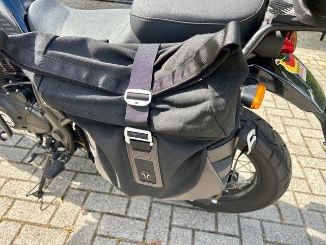 Schwarze Motorradsatteltasche, die an einem geparkten Roller auf einer gepflasterten Straße befestigt ist.