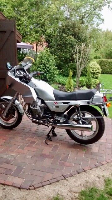 Ein silber-schwarzes Motorrad parkt auf einer gepflasterten Auffahrt neben einem Garten mit Bäumen und Sträuchern.