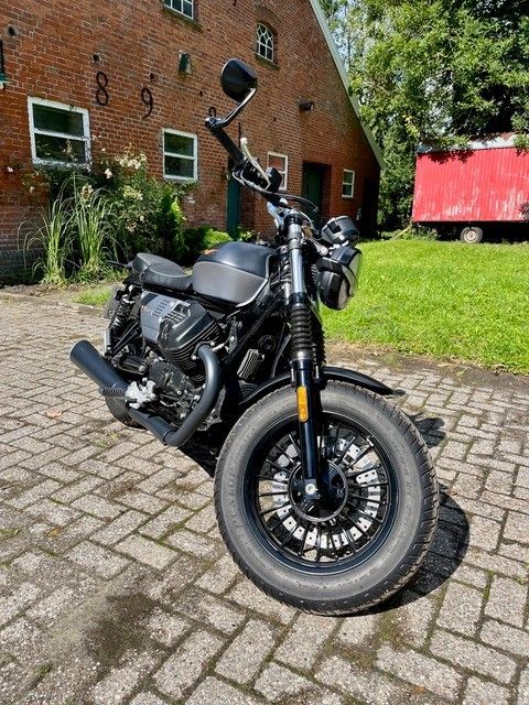 Schwarzes Motorrad, das auf Kopfsteinpflaster vor einem Backsteingebäude und grünem Rasen geparkt ist.