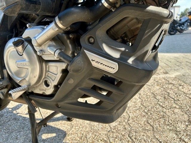 Nahaufnahme eines Motorradmotors und einer schwarzen Unterfahrschutzplatte auf einer gepflasterten Fläche.