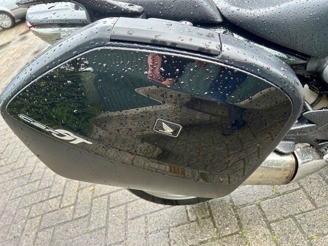 Nahaufnahme einer schwarzen Motorradseitenabdeckung mit Regentropfen, die auf nassem Gehweg geparkt ist.