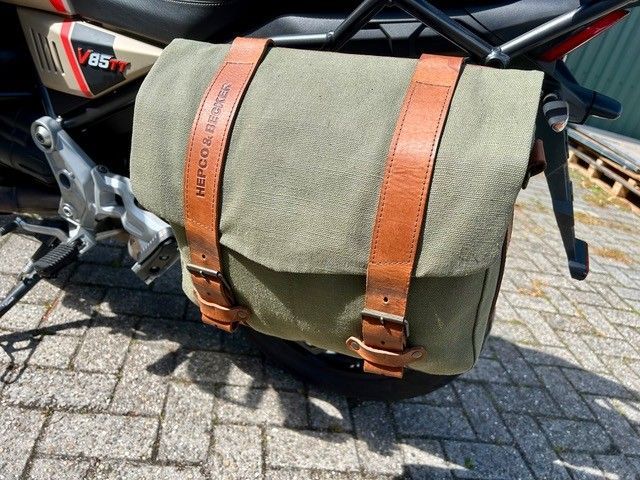 Olivgrüne Motorradsatteltasche mit braunen Riemen auf einem geparkten Motorrad über Backsteinpflaster