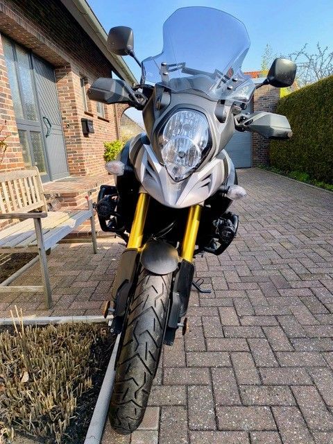 Ein graues Suzuki V-Strom-Motorrad mit gelben Gabeln, geparkt auf einer gepflasterten Auffahrt vor einem Haus.
