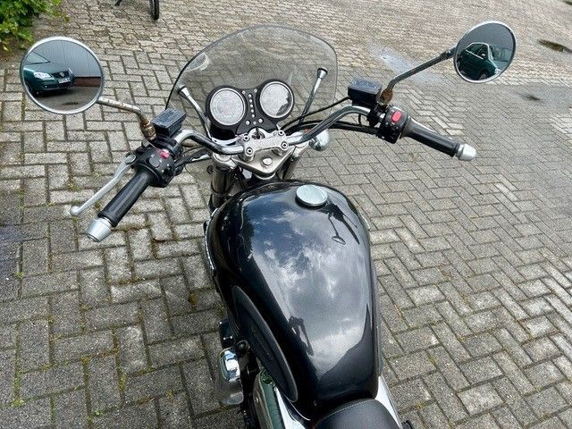Schwarzes Motorrad-Armaturenbrett und Lenker, von oben auf einer Kopfsteinpflasterstraße gesehen.