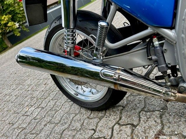 Verchromtes Motorradauspuffrohr mit Rad, Stoßdämpfern und blauer Karosserie in einer grau gepflasterten Umgebung.