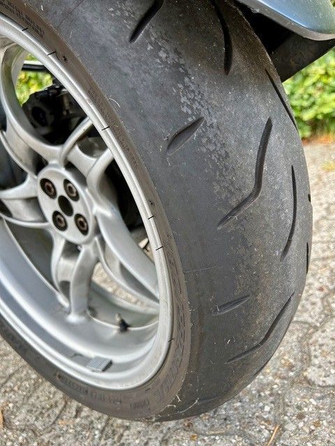 Nahaufnahme eines Motorradreifens auf einer silbernen Felge. Der Reifen ist abgenutzt, mit sichtbarem Profil und einigen Verfärbungen.